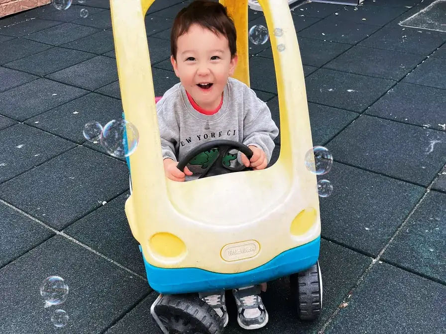 Laughing toddler riding a ride-on toy car among soap bubbles in the Richmond outdoor play yard.