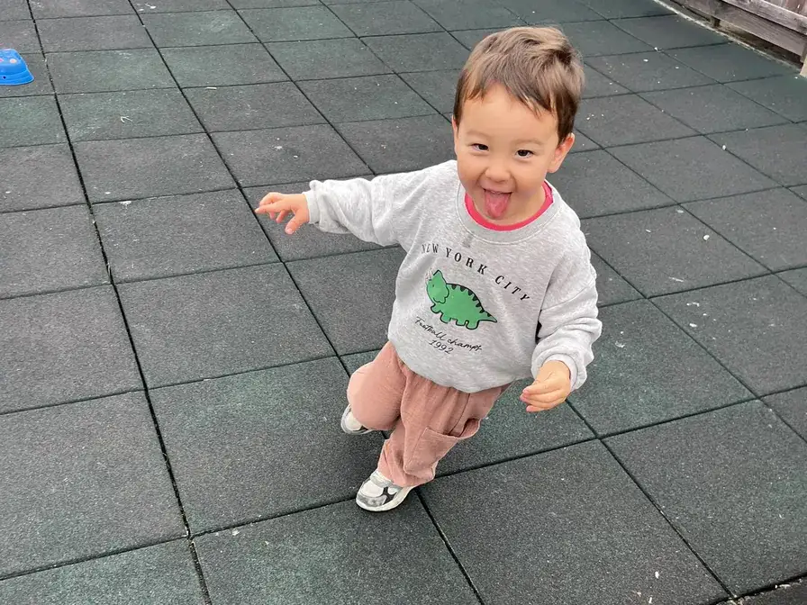 Toddler in a dinosaur sweatshirt running across the Richmond outdoor play yard with arms out.