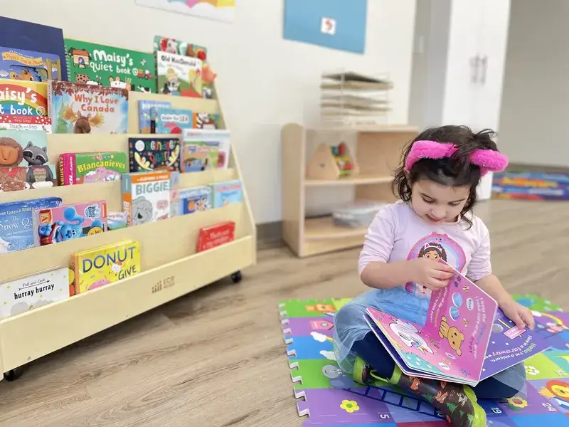 Child reading a picture book on a play mat beside the classroom bookshelf.