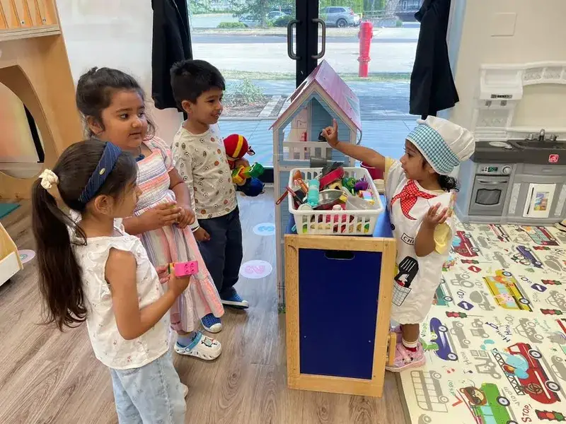 Child in a chef hat serving friends at the classroom pretend-play kitchen.