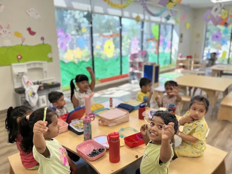 Smiling children holding up their snacks around a shared classroom lunch table.