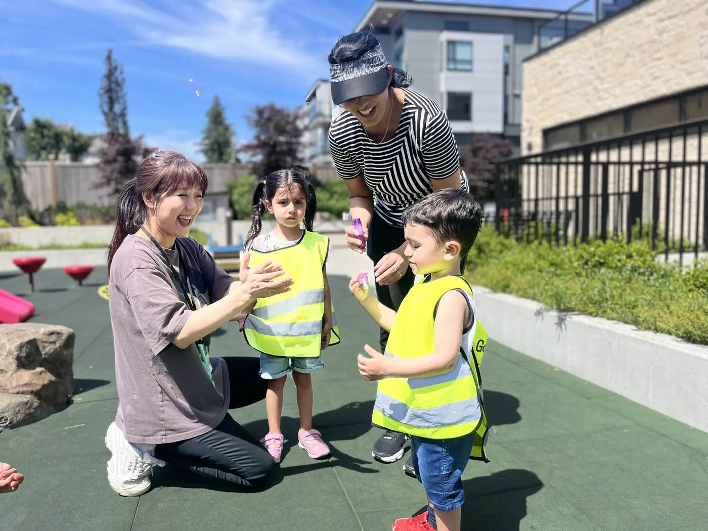 Educators blowing bubbles with two children in safety vests on a sunny outdoor playground.
